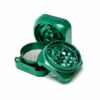 Open Krush 3.0 grinder in emerald green, showing the square lid, sharp teeth, mesh screen and lower chamber on a light grey background.